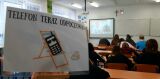 Na pierwszym planie tabliczka z rysunkiem telefonu na leżaku i napisem 'TELEFON TERAZ ODPOCZYWA :)', w tle uczniowie w klasie.