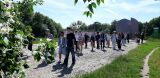 Grupa młodzieży i opiekunów spaceruje brukowaną aleją w stronę nowoczesnego pomnika w słoneczny dzień.