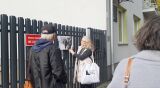 Edukacyjny spacer: przewodniczka pokazuje uczestnikom czarno-białą fotografię historyczną przy ogrodzeniu budynku.