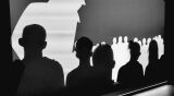 Zbliżenie na sylwetki widzów na tle jasnego ekranu z konturami wielu małych postaci i jedną dużą figurą.