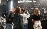 Uczniowie piszą kredą na dużej, czarnej tablicy z nagłówkiem 'dzieciństwo to…', która jest już gęsto pokryta napisami.