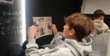 Chłopiec w szarej bluzie siedzi w pufie i z bliska czyta zadrukowany arkusz papieru ze zdjęciami.