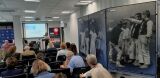 Sala konferencyjna z publicznością słuchającą prelegentki przy mównicy i ekranem projekcyjnym w tle.