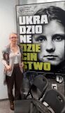 Kobieta w okularach i wzorzystym szalu stoi obok dużego plakatu z twarzą dziecka i napisem IPN.