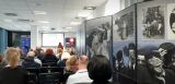 Sala konferencyjna z publicznością, prelegentką przy mównicy i dużymi panelami wystawowymi ze zdjęciami dzieci.