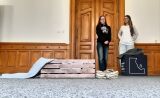 Dwie dziewczyny stojące przy drewnianych drzwiach obok rekwizytów w postaci drewnianej skrzyni, koca i zniszczonych butów.
