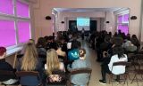 Widok z tyłu na uczniów siedzących w sali i słuchających prelekcji dwojga prowadzących.