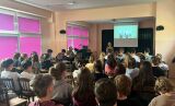 Sala lekcyjna wypełniona młodzieżą, która słucha kobiety przemawiającej na środku przed ekranem.