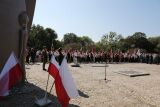 Delegacje z pocztami sztandarowymi stoją przy pomniku i polskich flagach na plenerowej ceremonii.