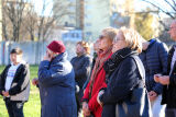 Uczestnicy uroczystości, głównie osoby starsze i dorośli, stoją w skupieniu pod gołym niebem, w jesiennej scenerii z blokami w tle.