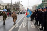 Grupa oficjeli i służb mundurowych stojących na jezdni podczas uroczystego przemarszu, na pierwszym planie salutujący żołnierz.