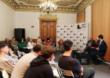 Uczestnicy wydarzenia siedzą na krzesłach i słuchają dyskusji prowadzonej przez panelistów w sali konferencyjnej. 
