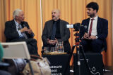 Starszy mężczyzna przemawia do mikrofonu podczas panelu, siedząc obok dwóch innych uczestników. 