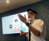 Bliskie ujęcie brodatego mężczyzny w okularach i czapce, który przemawia, wskazując na ekran z wyświetloną aplikacją edukacyjną Kialo.