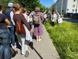 Grupa osób idzie chodnikiem wzdłuż trawnika, podążając za przewodniczką niosącą materiałową torbę.