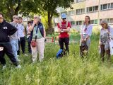 Grupa dorosłych i młodzieży stoi w wysokiej trawie przed blokiem mieszkalnym, słuchając kobiety trzymającej historyczną fotografię.