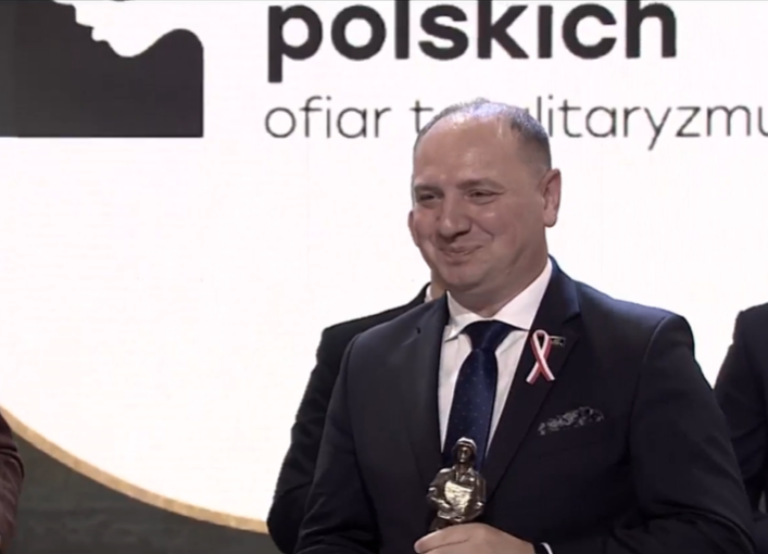 Mężczyzna z nagrodą, uśmiechający się na tle plakatu.