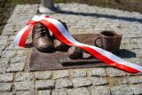 Pomnik z butami, kubkiem i wstęgą narodową na kostce brukowej.