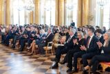Rzędy gości w garniturach i eleganckich strojach siedzących na krzesłach i bijących brawo.