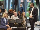 Mężczyzna w garniturze przemawia do mikrofonu przed grupą siedzących osób w nowoczesnej sali.