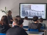 Zbliżenie na uczniów patrzących na archiwalną fotografię wyświetloną na ekranie, przedstawiającą grupę więźniów w pasiakach stojących w szeregu.