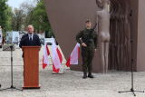 Mężczyzna przy mównicy, żołnierz w mundurze obok flag Polski.