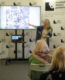 Kobieta wskazuje na ekranie archiwalne zdjęcie lotnicze terenu obozowego podczas prelekcji.