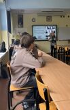 Prelegentka prowadzi wykład w klasie szkolnej przed ekranem z prezentacją o tytule 'Muzeum Dzieci Polskich', a uczniowie słuchają jej w ławkach.