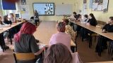 Kobieta przemawia do uczniów siedzących w ławkach w klasie szkolnej, prezentując slajd na ekranie multimedialnym.