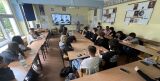 Klasa szkolna z lotu ptaka, w której uczniowie siedzą przy złączonych stołach, a nauczycielka wskazuje coś na ekranie multimedialnym.