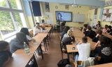 Widok na klasę lekcyjną z perspektywy tylnej ławki, w której młodzież uczestniczy w prelekcji historycznej prowadzonej przez kobietę.