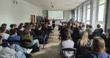Prelekcja historyczna w pełnej sali wykładowej, gdzie słuchacze siedzą przodem do ekranu z prezentacją i stojących prelegentów.