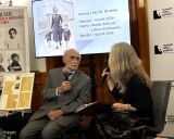 Bohdan Kończak z mikrofonem siedzi obok prowadzącej warsztaty na tle ekranu wyświetlającego archiwalne zdjęcie kobiety z dziećmi.