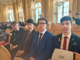 Czterech młodych mężczyzn w garniturach siedzi w rzędzie w zabytkowej, złoconej sali. Jeden z nich trzyma białą kartkę z czarnym napisem 'VIP'.