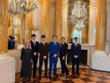 Grupa sześciu osób w eleganckich strojach pozuje w bogato zdobionej sali balowej z dużymi żyrandolami i lustrami.